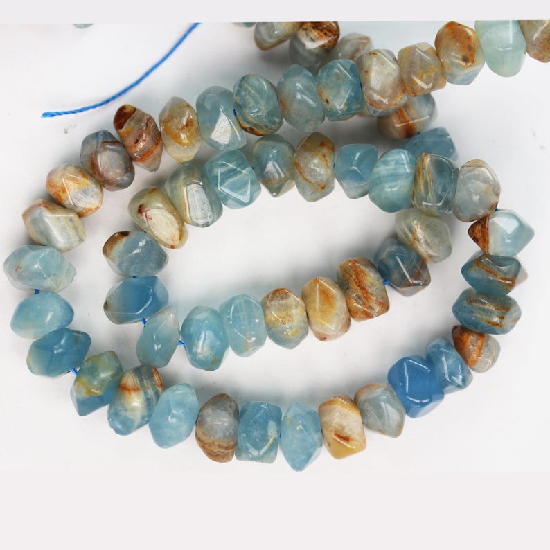 May include: A strand of faceted, irregular gemstone beads in shades of light blue and translucent white with streaks of warm brown. The beads are strung on a thin blue thread, displaying a polished, multi-faceted surface texture throughout.