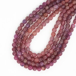 May include: A strand of pink and purple gemstone beads. The beads are round and have a smooth, polished finish. The beads are strung on a red thread.