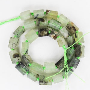 Op de afbeelding: Een streng groene en zwarte edelsteen kralen, elke kraal heeft de vorm van een rechthoekig prisma. De kralen zijn geregen op een groene draad.