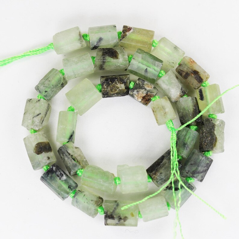 Op de afbeelding: Een streng groene en zwarte edelsteen kralen, elke kraal heeft de vorm van een rechthoekig prisma. De kralen zijn geregen op een groene draad.