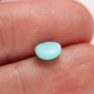 May include: A small, oval-shaped, polished gemstone in a light turquoise color. The stone is smooth and appears to be held in a person's hand. The gemstone is a cabochon cut, with a rounded top and a flat bottom.