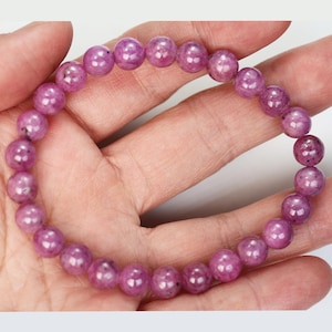 May include: A bracelet of round, polished, pink gemstones. The beads are strung together, forming a complete circle. The bracelet is held in a hand, showcasing the colour and texture of the stones. The gemstones have a slight translucence.