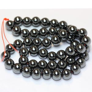 Peut inclure: Un brin de perles d'hématite noire, chaque perle est ronde et lisse avec un poli élevé. Les perles sont enfilées sur un fil rouge.