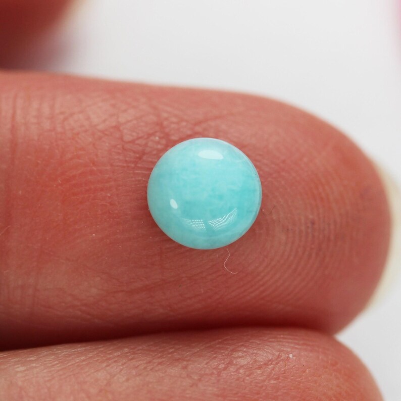 May include: A round, polished, light blue gemstone. The gemstone is a cabochon cut, with a smooth, domed surface. The color is a soft, pastel blue, with a slight translucence. The gemstone is likely a bead or a small decorative element.
