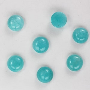 May include: Seven round, polished, turquoise-colored gemstones are arranged against a white background. The stones have a smooth, glossy surface, reflecting light. The gemstones are a vibrant, solid color, and appear to be cabochons, suitable for jewelry making or crafts.