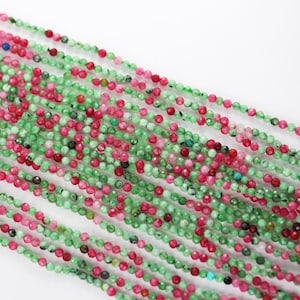 Op de afbeelding: Een streng groene en roze edelsteen kralen. De kralen zijn klein en rond en geregen aan een dunne draad. De kralen zijn een mix van groen en roze en hebben een licht onregelmatige vorm.