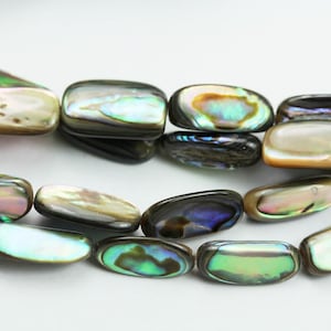 May include: Close-up of iridescent abalone shell beads. The beads are rectangular with rounded edges, displaying a range of colors including green, blue, and gold. The beads are strung together, suggesting they are for jewelry making.