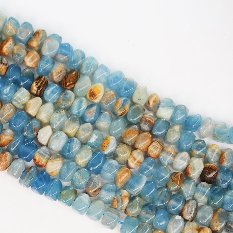 May include: A strand of tumbled gemstone beads featuring a mix of translucent light blue and opaque tan or brown marbled patterns. Each bead has an irregular, rounded shape with a smooth, polished finish, arranged in multiple parallel rows.