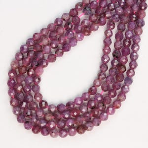 Puede incluir: Un hilo de cuentas de piedras preciosas facetadas, redondas, de color rosa oscuro y morado, ensartadas en un cordón rosa fino. Las cuentas muestran una calidad translúcida con inclusiones naturales y una textura superficial multifacetada.