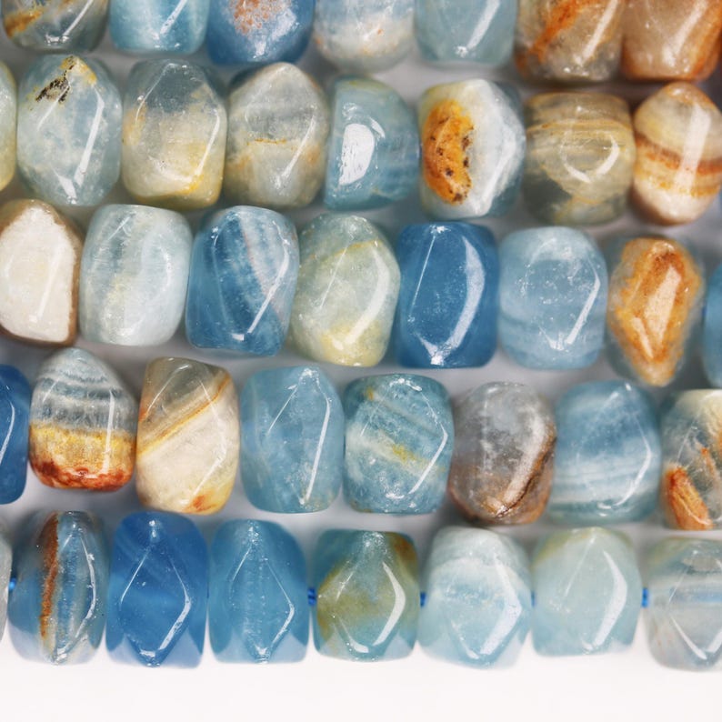 May include: A collection of tumbled, rectangular-shaped beads featuring a translucent light blue base with visible internal banding and inclusions in shades of white, cream, and golden brown, arranged in a grid-like pattern on a white surface.