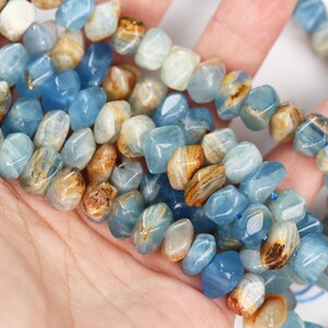 May include: A strand of tumbled gemstone beads featuring a mix of translucent light blue, opaque white, and earthy brown tones. The beads have an irregular, faceted shape and are strung together on a thin blue cord, showcasing natural color variations.