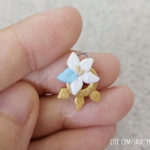 Puede incluir: Un amuleto dorado con una flor blanca y un centro azul, con un lazo plateado para sujetarlo a un collar o pulsera.
