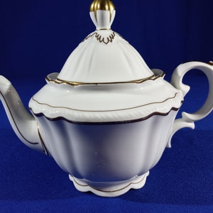 May include: White porcelain teapot with a gold trim and a lid. The teapot has a fluted design and a scalloped edge.