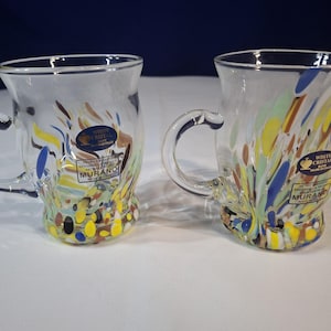 May include: Two clear glass mugs with handles, adorned with colorful abstract designs. The mugs feature a base of yellow, brown, and blue speckles, with streaks of color ascending the sides. Each mug has a label that reads "WHITE CRISTAL MADE IN ITALY".