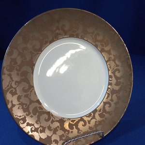 May include: A large, round, white ceramic dinner plate with a gold rim and a swirling, embossed pattern of clouds. The plate is on a blue background.