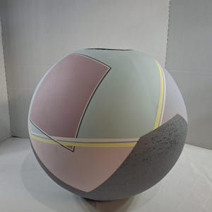 May include: A spherical ceramic vase featuring a matte finish with geometric color blocking in shades of dusty rose, sage green, and pale blue. The design includes thin yellow and white accent lines and a textured dark gray speckled base section.