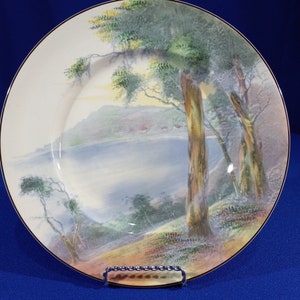 May include: A vintage ceramic plate with a scenic landscape design. The plate features a watercolor-style painting of trees, a lake, and a distant hillside. The plate has a gold rim and a white background.