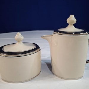 May include: A cream-colored ceramic sugar bowl and creamer set featuring a smooth finish, silver-toned metallic trim along the rims and handle, and matching lids topped with small, pointed, bud-shaped finials for a cohesive appearance.