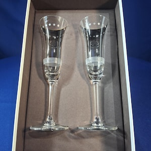 May include: Two clear champagne flutes with white dot and stripe detailing, presented in a brown box. The flutes have a slender stem and a wide, flared bowl. The rim of the flutes has a gold trim.