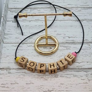 May include: A wooden name necklace with the name "Sophie" spelled out in individual blocks. The necklace is on a black cord and has pink and yellow beads.