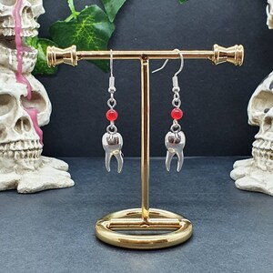 Könnte beinhalten: Silberfarbene, zahnförmige Ohrringe mit roten Perlen, die an silbernen Haken hängen. Die Ohrringe werden auf einem goldfarbenen Ständer vor dunklem Hintergrund präsentiert. Ein einzigartiges Accessoire.