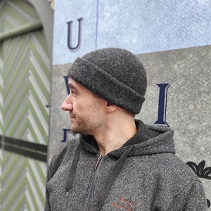 Puede incluir: Una persona con una sudadera con capucha gris y una boina gris. La persona está de pie frente a una pared con la letra "U" pintada en ella.