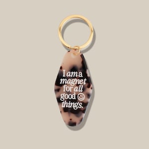 Può includere: Un portachiavi con un anello dorato e un'etichetta fantasia. L'etichetta presenta un motivo tartarugato beige e marrone e il testo "I am a magnet for all good things." con una faccina sorridente.