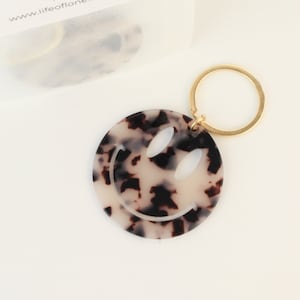 Può includere: Un portachiavi rotondo con un design a faccina sorridente. Il portachiavi ha un motivo a scaglie di tartaruga nei toni del crema e del marrone. È dotato di un portachiavi in metallo color oro. L'indirizzo del sito web "www.lifeoflone.com" è visibile sullo sfondo.