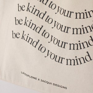 May include: A white canvas tote bag with the text "be kind to your mind" repeated four times in a curved pattern. The text is in black and the bag has the text "LIFEOFLONE X JACQUI DESIGNS" printed on the bottom.