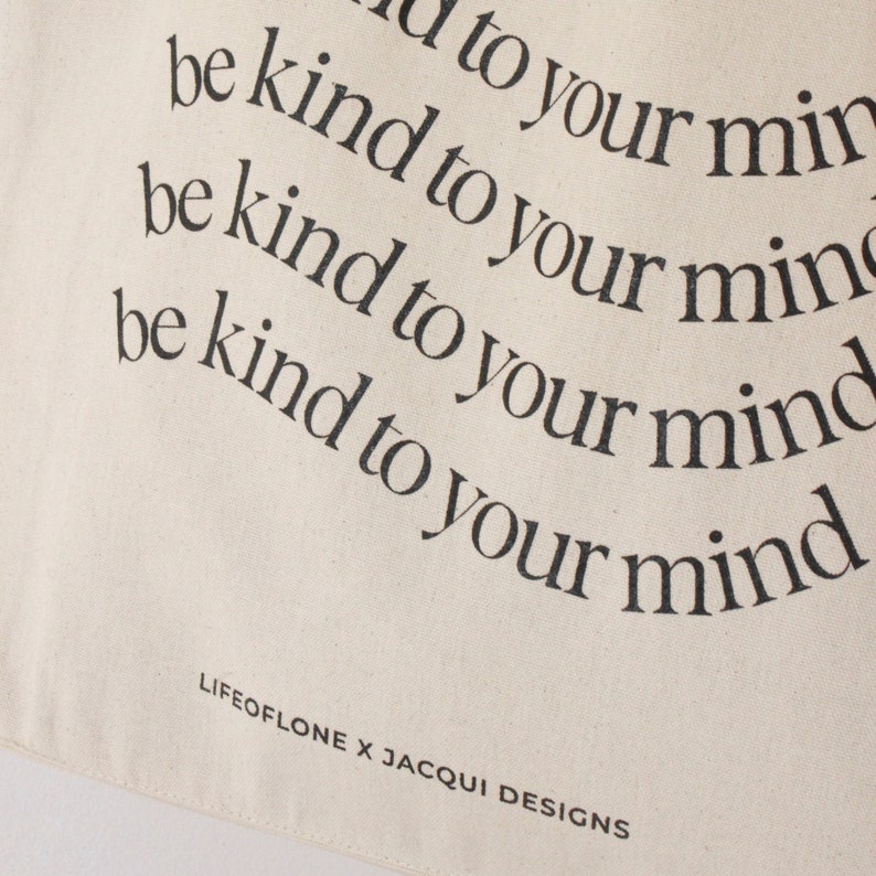 May include: A white canvas tote bag with the text "be kind to your mind" repeated four times in a curved pattern. The text is in black and the bag has the text "LIFEOFLONE X JACQUI DESIGNS" printed on the bottom.
