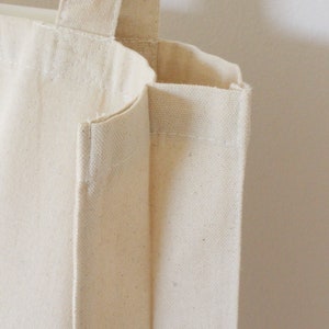May include: A close-up of a plain, off-white canvas tote bag with a single handle. The bag is made of a thick, durable fabric and has a simple, classic design.
