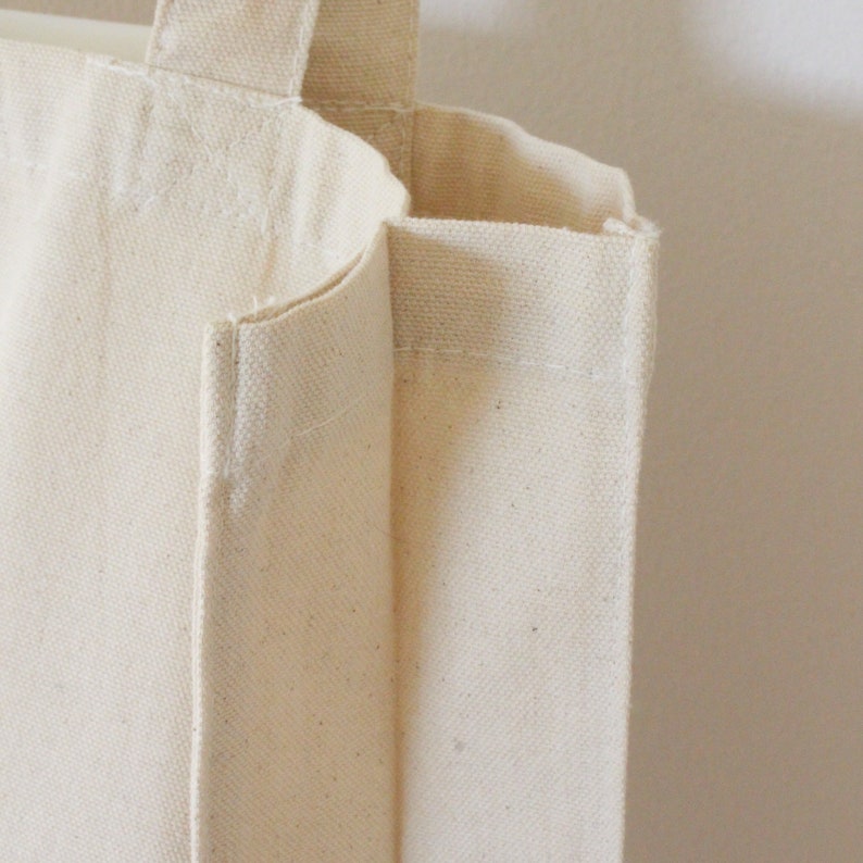 May include: A close-up of a plain, off-white canvas tote bag with a single handle. The bag is made of a thick, durable fabric and has a simple, classic design.