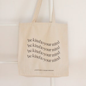 May include: A beige canvas tote bag with the text "be kind to your mind" repeated four times in a circular pattern. The text is in black and the bag has black handles. The bottom of the bag has the text "LIFEOFLONE X JACQUI DESIGNS" in black.