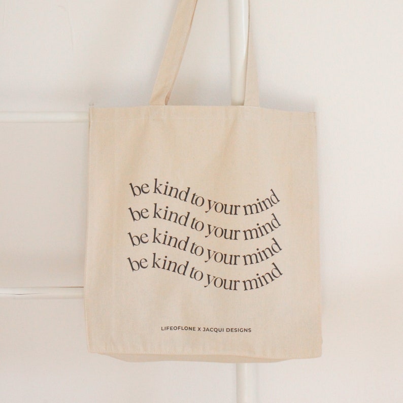 May include: A beige canvas tote bag with the text "be kind to your mind" repeated four times in a circular pattern. The text is in black and the bag has black handles. The bottom of the bag has the text "LIFEOFLONE X JACQUI DESIGNS" in black.