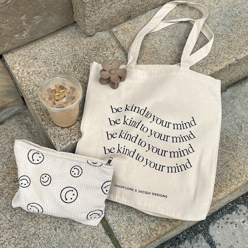 May include: A beige canvas tote bag with the text "be kind to your mind" repeated four times. The bag has brown straps and a brown flower hair clip attached to the strap. A small beige pouch with a white smiley face pattern is in the foreground.