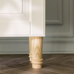 Könnte beinhalten: Nahaufnahme eines weißen Schranks mit Holzbeinen. Der Schrank steht auf einem Holzboden.