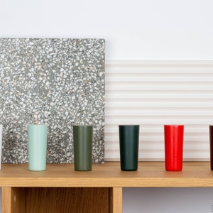 Op de afbeelding: Zeven hoge, cilindrische glazen in verschillende kleuren, waaronder wit, groen, donkergroen, donkerblauw, rood, bruin en zwart, staan op een houten plank.