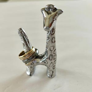 May include: A silver giraffe ring holder with a textured surface. Three rings are on the holder, one gold and two silver.