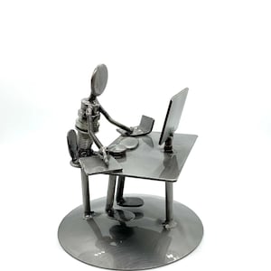 May include: A metal sculpture of a person at a desk with a computer and laptop. The figure is made of metal pieces welded together, with a round base. The desk has a monitor, laptop, and other small items.