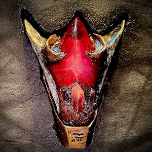 May include: A red crystal with gold accents in the shape of a stylized skull. The crystal is set in a gold frame with a decorative pattern.