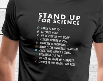May include: Black t-shirt with white text that reads "Stand Up For Science" and a list of scientific facts. The list includes: Earth is not flat, vaccines work, we've been to the moon, climate change is real, universe is expanding, math is the universal language, chemtrails aren't a thing, evolution is a fact, we are all made of stardust, science is like magic, but real.