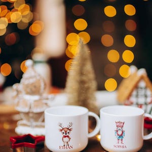 May include: Two white ceramic mugs with holiday designs. One mug features a reindeer with the name "ETHAN", the other a nutcracker with the name "SOPHIA". Festive Christmas decor includes a star cookie cutter and a small gold tree.