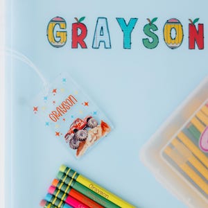 May include: A light blue binder with the name "Grayson" in colorful letters. A clear tag with the name "Grayson" and a monster truck graphic hangs from the binder. Several pencils with the name "Grayson" are also visible.