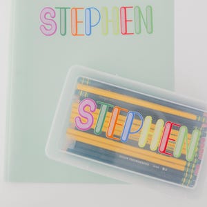 Puede incluir: Un archivador y un estuche de lápices verde claro con el nombre "STEPHEN" en letras coloridas. El estuche está lleno de lápices amarillos.