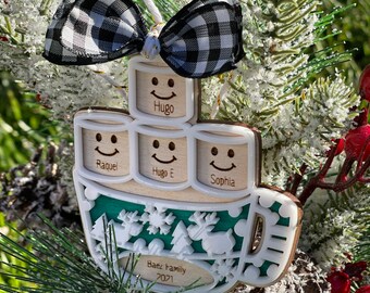 May include: A wooden Christmas ornament shaped like a mug with four marshmallows. Each marshmallow has a smiley face and a name: Hugo, Raquel, Hugo E, and Sophia. The ornament is decorated with a green and white snowflake pattern and the text "Baez Family 2021".
