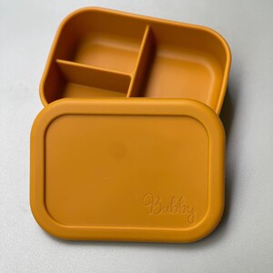 May include: A mustard yellow silicone lunch box with a divided interior and a lid that has the word "Bubbly" embossed on it.