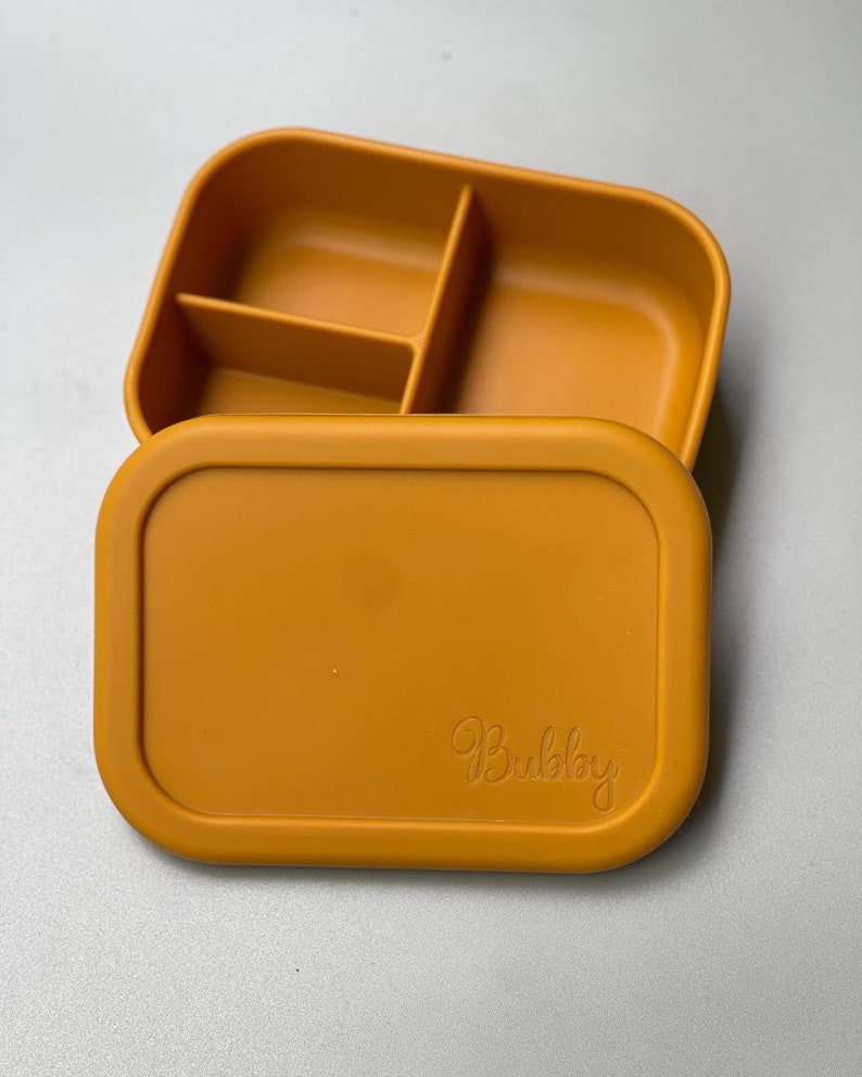 May include: A mustard yellow silicone lunch box with a divided interior and a lid that has the word "Bubbly" embossed on it.