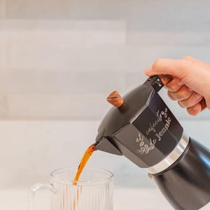 May include: A black stovetop espresso maker with a wooden handle and lid is pouring coffee into a clear glass mug. The espresso maker has the text "El cafecito de Jennie" on the side.