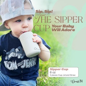 May include: A baby is drinking from a light gray sipper cup. The cup has the word "Anso" printed on it. The baby is wearing a navy blue t-shirt with a Tom and Jerry graphic and blue denim shorts. The image includes the text "Sip, Sip! The Sipper Cup Your Baby Will Adore."