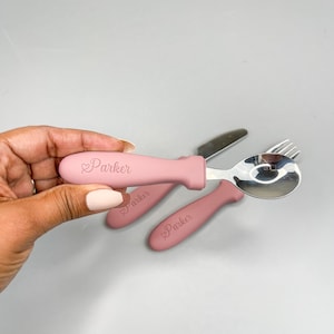 May include: A set of three pink baby utensils with a silicone handle. The utensils are a fork, spoon, and knife. The handle has the name "Parker" engraved on it.